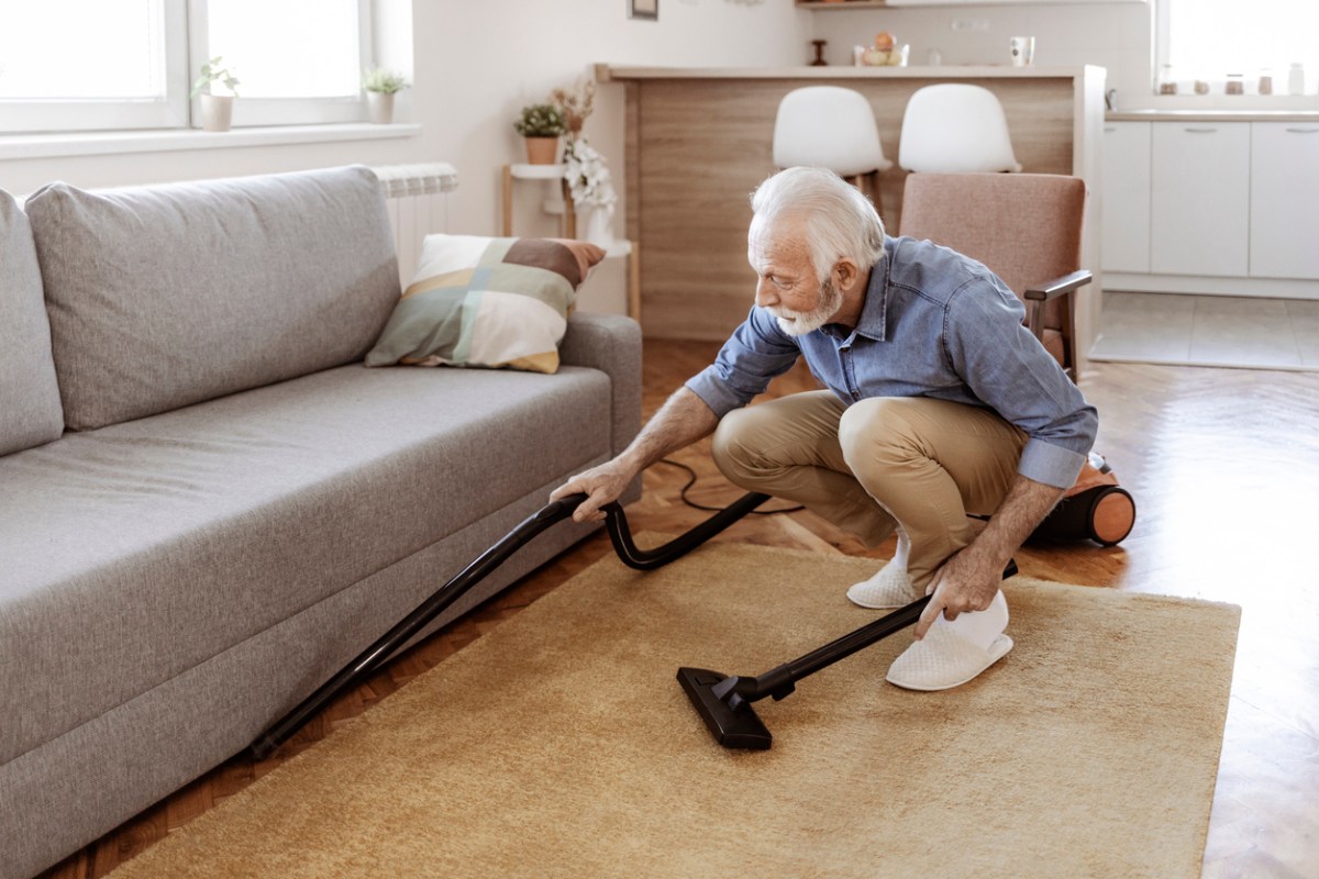 A senior crouched down using the best lightweight vacuum for seniors to clean under a couch.