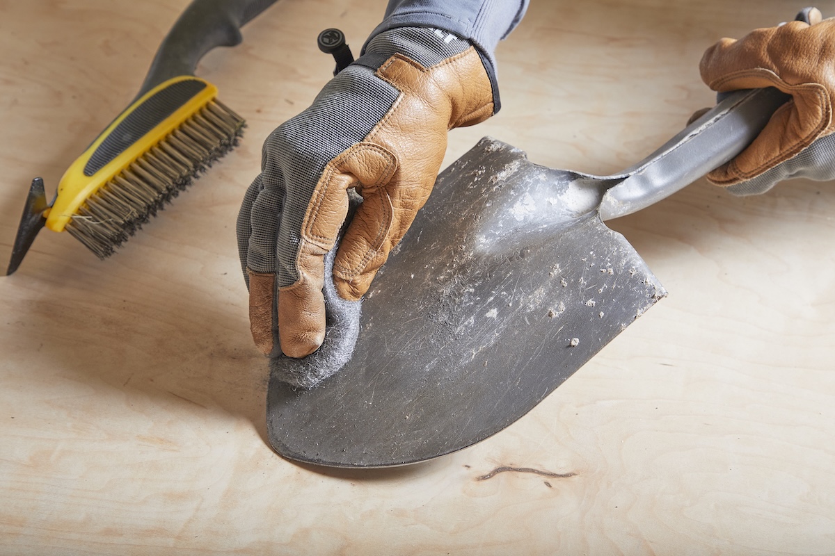 Person wearing work gloves uses steel wool to scrub the back of a garden shovel.