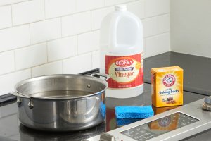 Large stainless pot, vinegar, baking soda and sponge on a stovetop.
