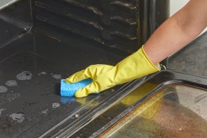Woman wearing rubber gloves wipes debris out of oven with blue sponge.
