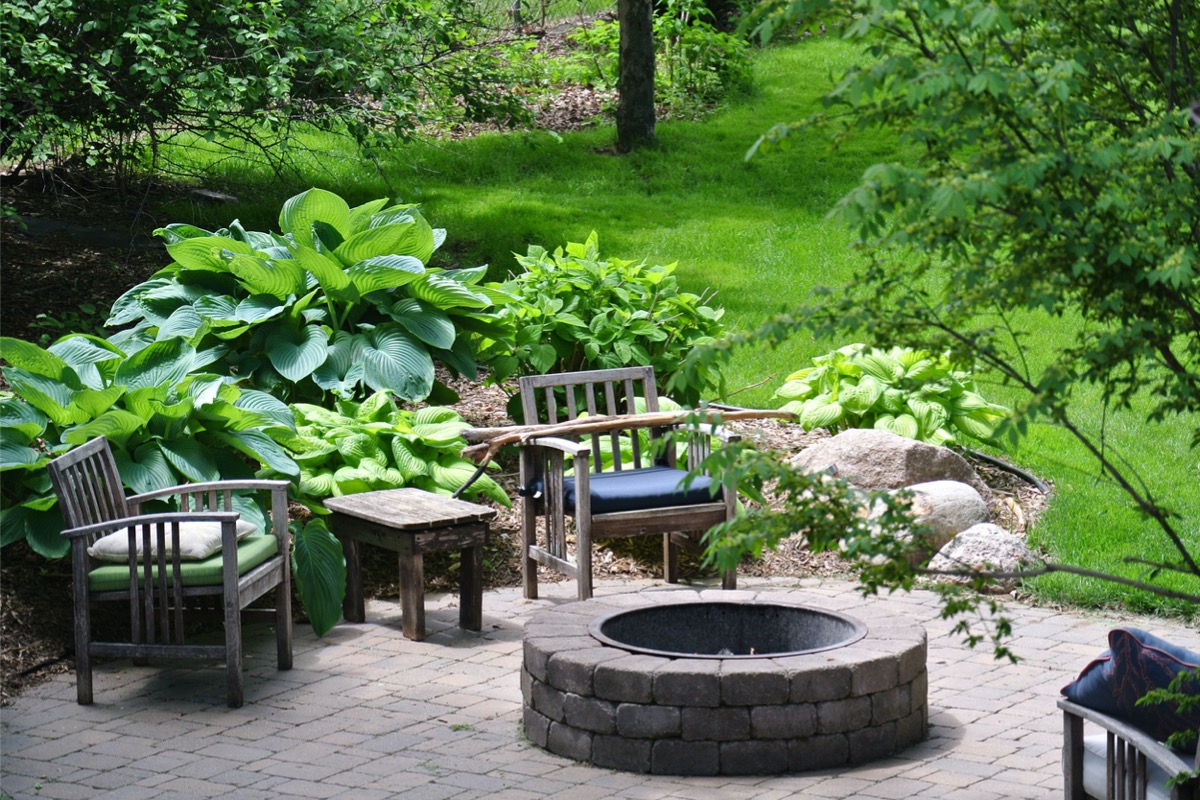 Lush back patio with green plantings, stone firepit, and stone patio.