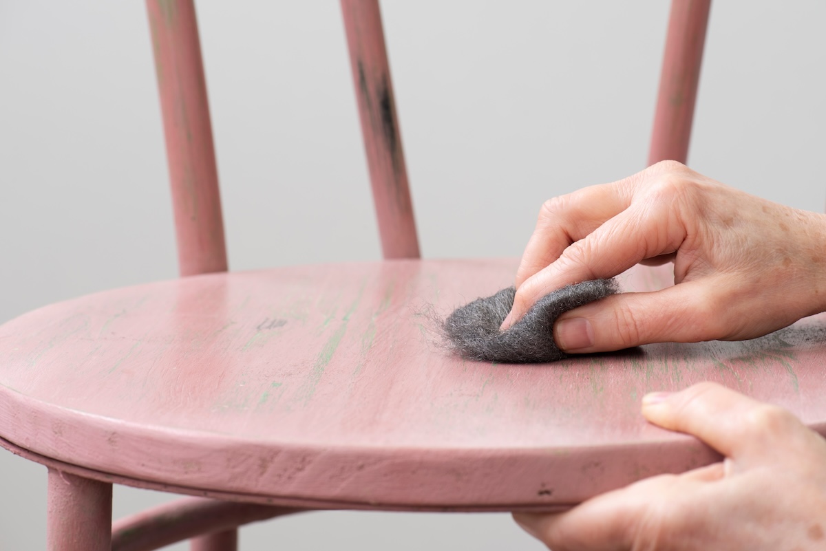 View of a person's hand using steel wool to sand the seat of a wooden chair painted pink.