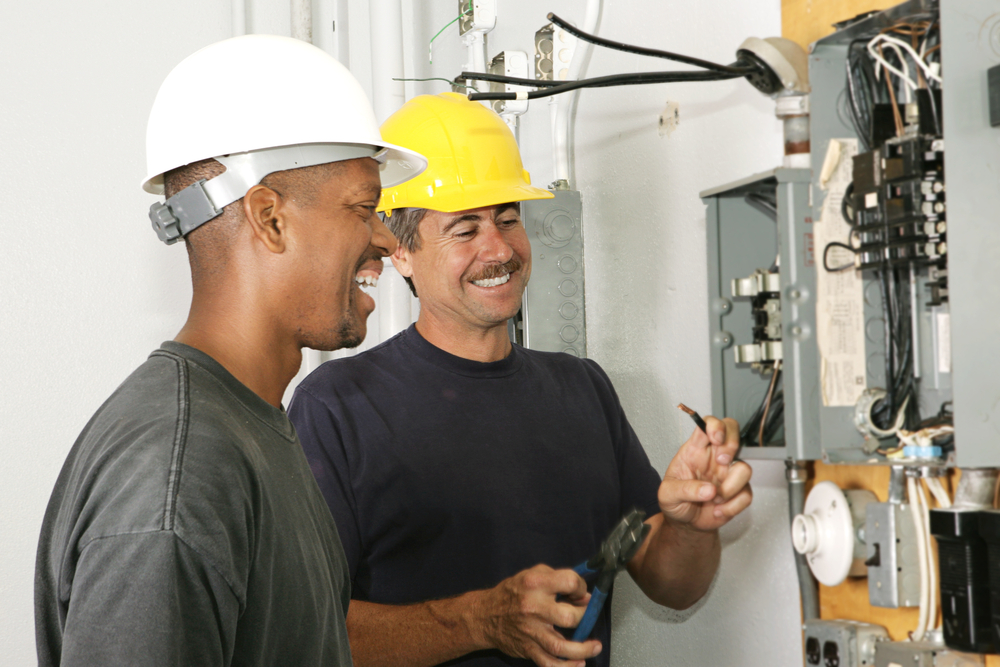 Two workers in hard hats smile and assess an electrical panel.