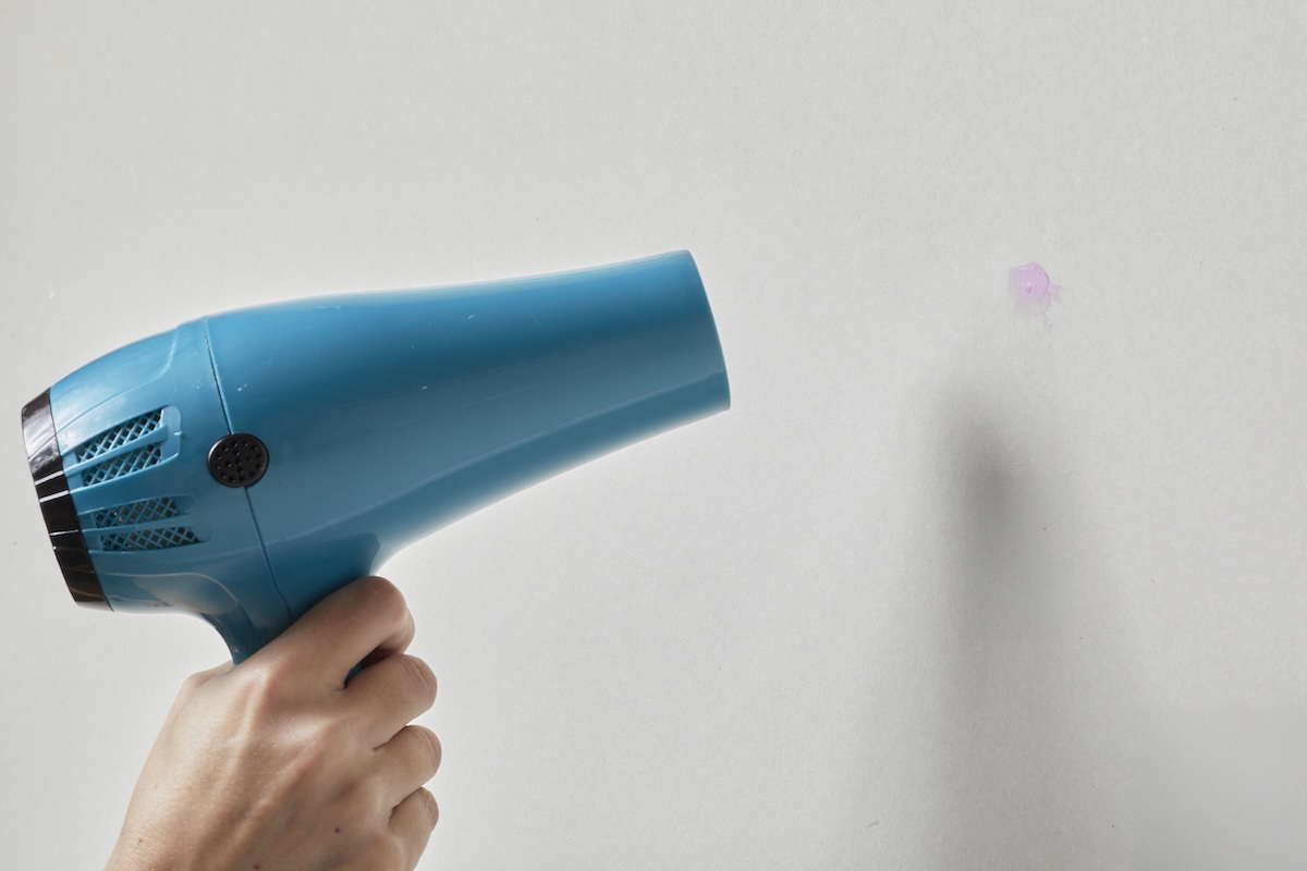 Person points hair dryer at hole in a wall covered with pink spackle.