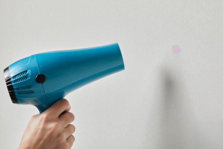 Person points hair dryer at hole in a wall covered with pink spackle.