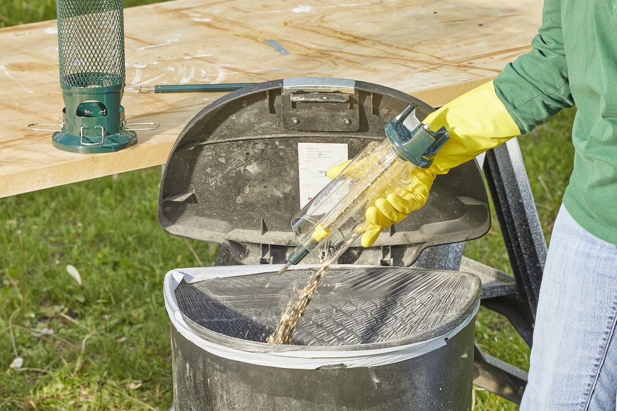 Woman wearing gloves dumps birdseed into a trash can.