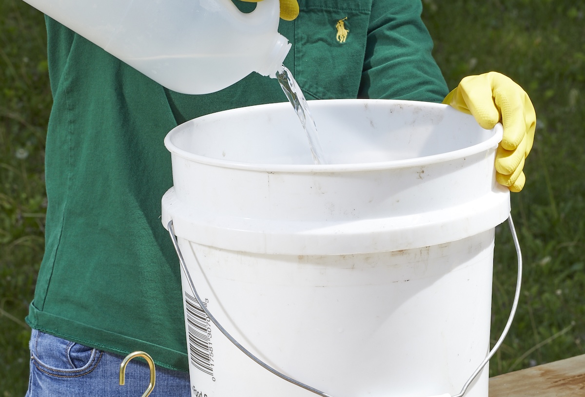 Woman pours vinegar from large jug into large white bucket.