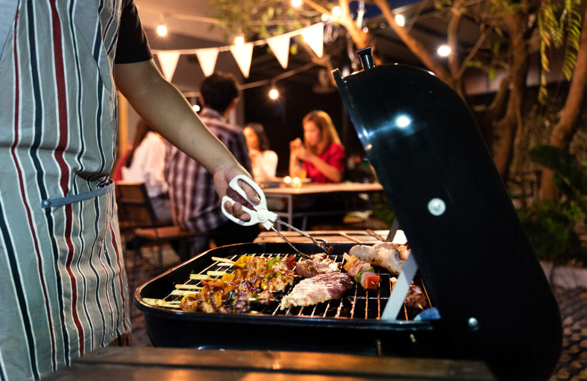 A person using tongs to flip food on a grill with a group sitting at a picnic table in the background