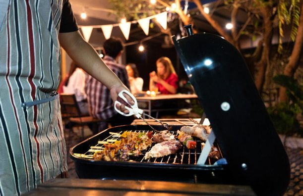 A person using tongs to flip food on a grill with a group sitting at a picnic table in the background
