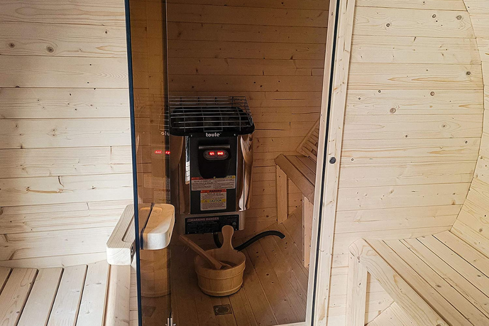 The Best Sauna Heater installed in a wood sauna next to a built-in bench with a water bucket and scoop on the floor.