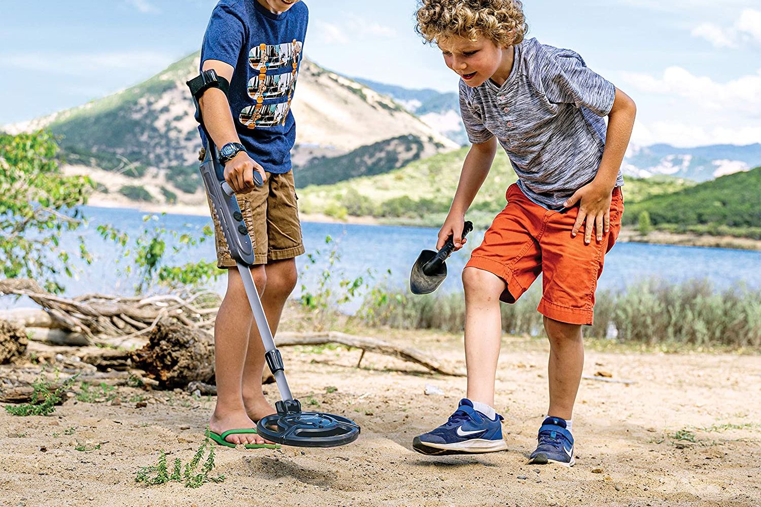 Two children using the best metal detector for beginners in the sand next to a mountain lake.