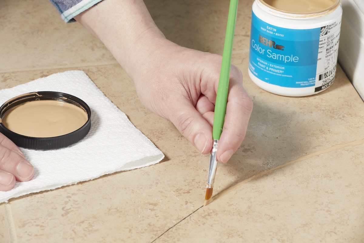 Woman uses detail paintbrrush to paint the crack of a cracked tile with beige paint.