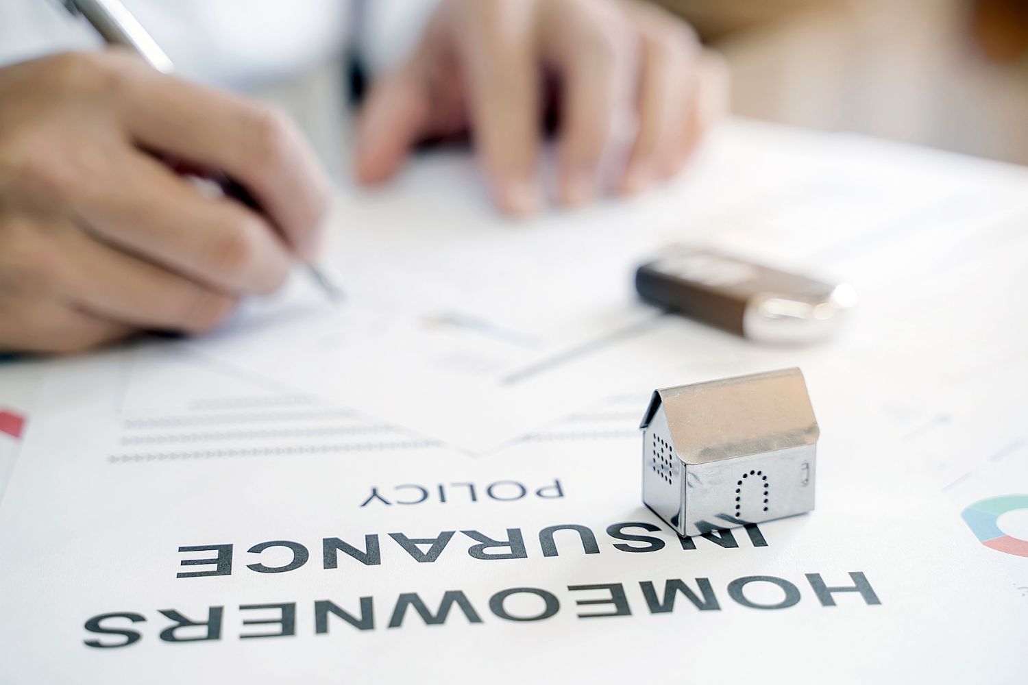 A small model house sits on a document called 'Homeowners insurance policy' while a person writes in the background.