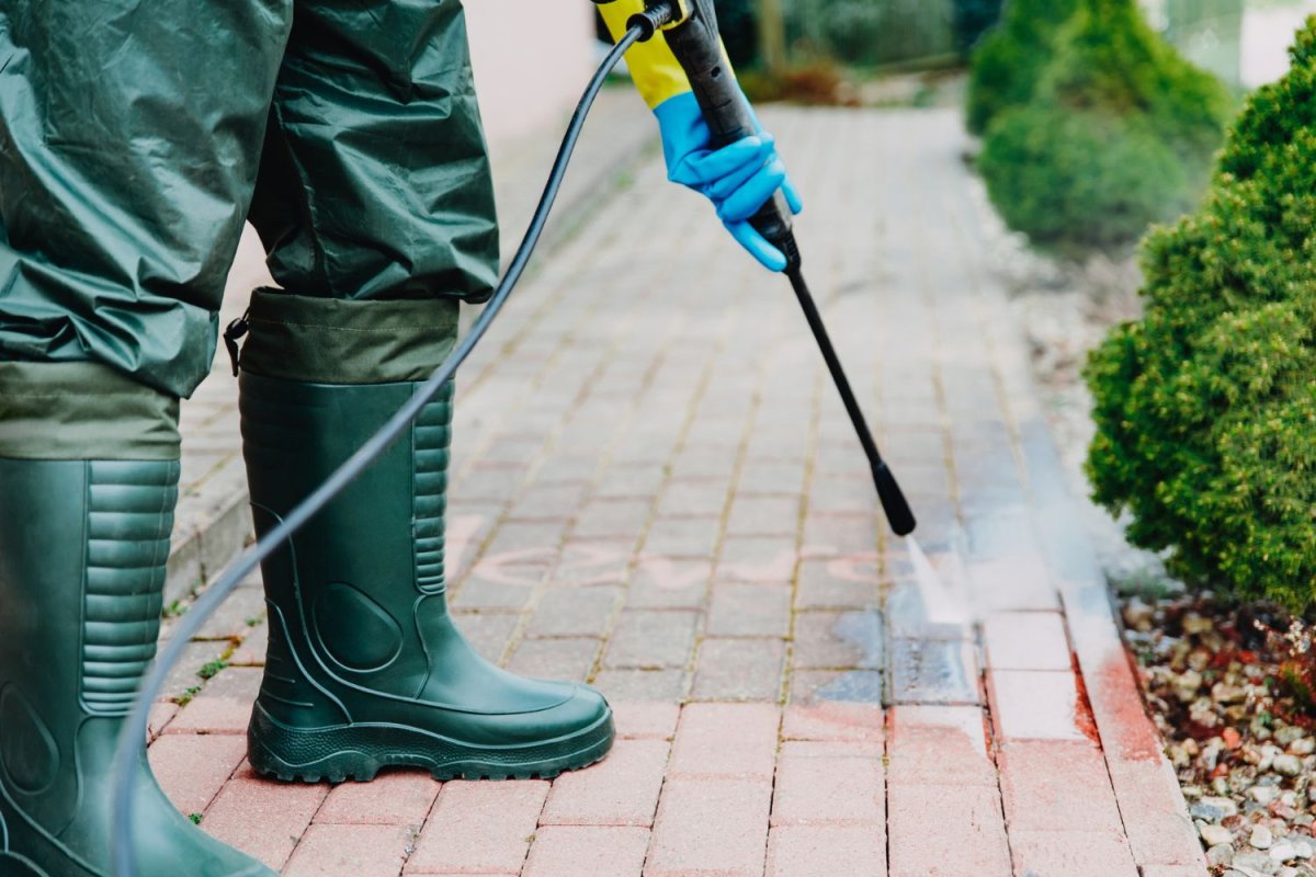 Person wearing tall rubber boots pressure washes a brick walkway.