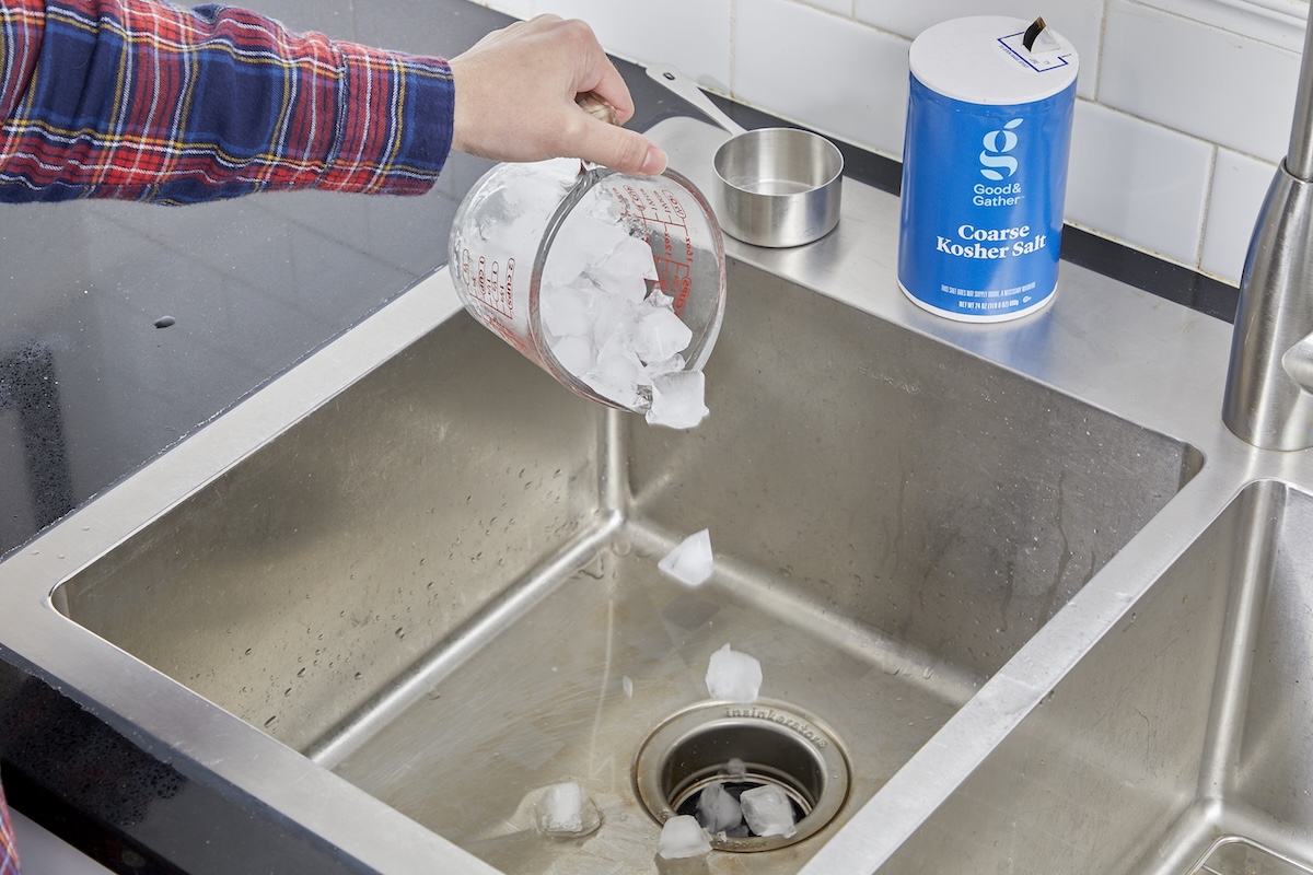 Woman pours measuring cup full of ice cubes over sink into garbage disposal, a container of salt resting nearby.
