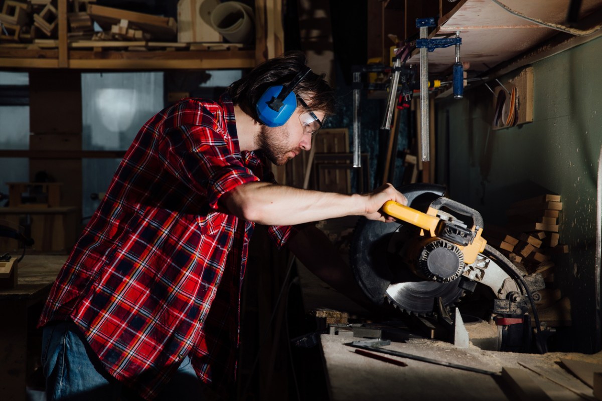 Man in flannel shirt wearing ear protection and using a circular saw in a workshop