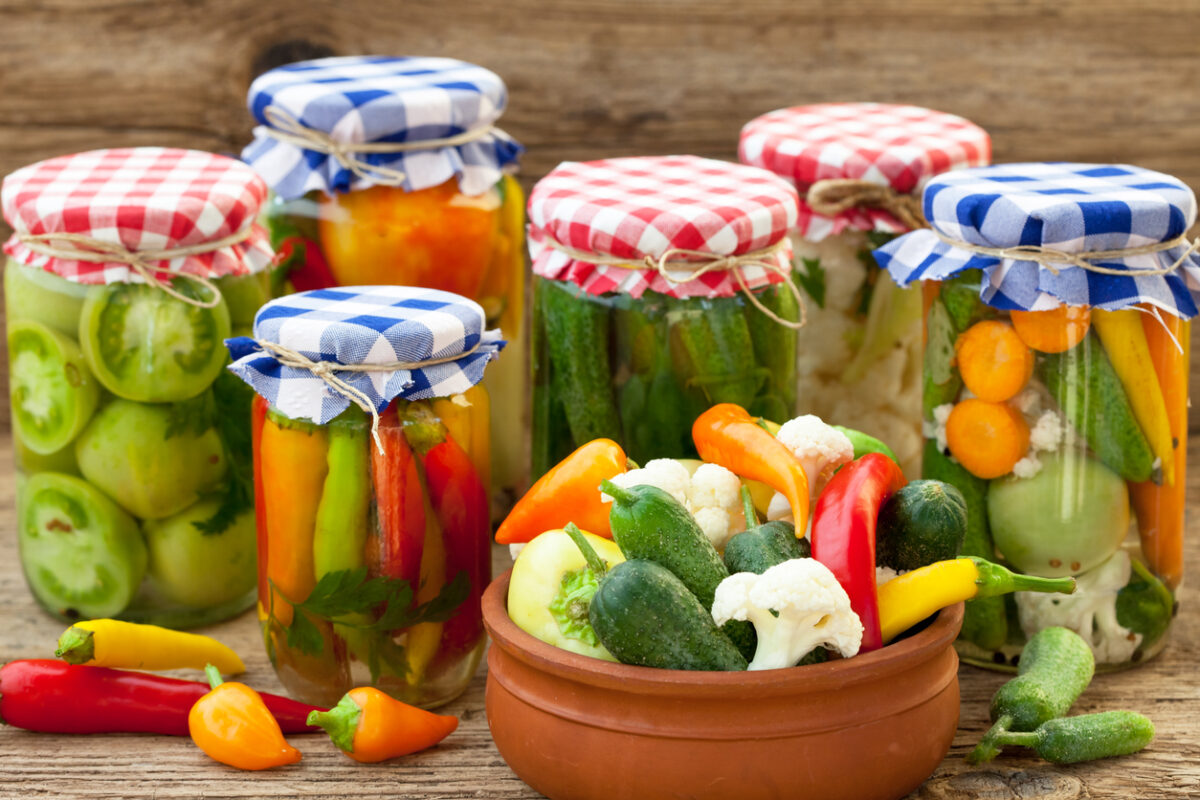 Glass jars with gingham-printed lids. The jars contain colorful vegetables. There's a bowl of fresh vegetables in the foreground.