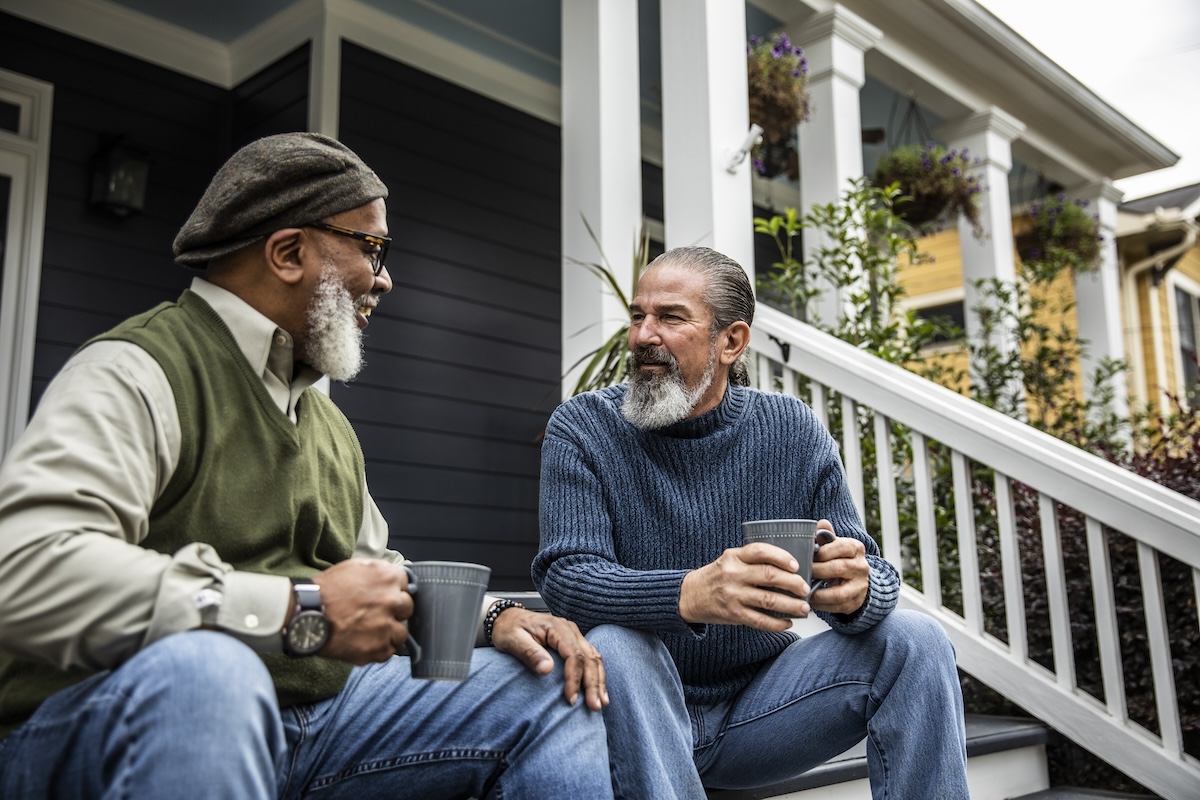Two senior men chatting on their front porch with coffee cups in hand.