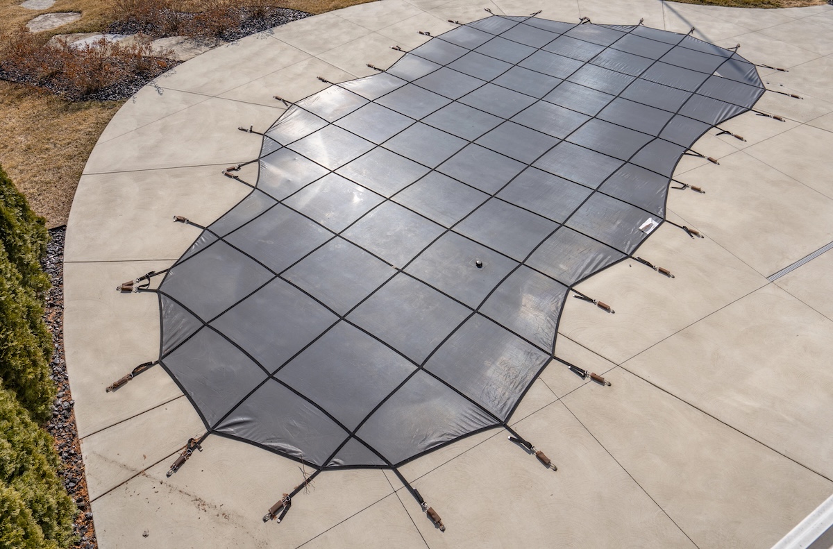 Kidney-shaped inground pool that has a cover over it with criss-cross ropes over it.