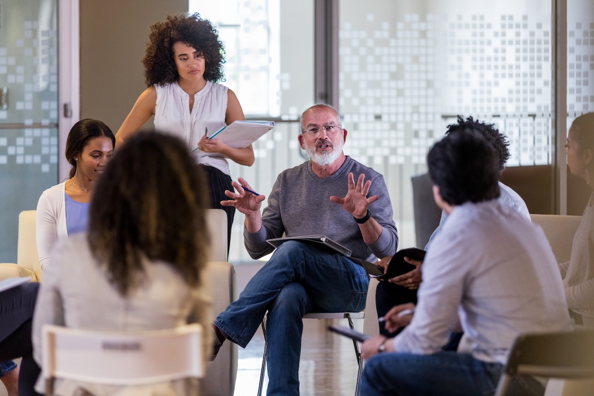 A group of people listening to an older man with a beard present at a Home Owners Association meeting.