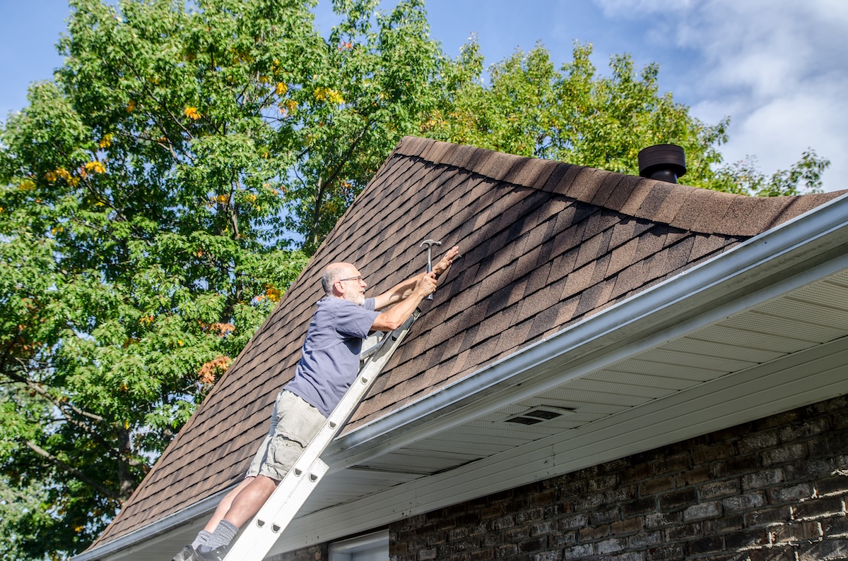 Middle-aged man standing on ladder and repairing an asphalt roof shingle near the roof peak.