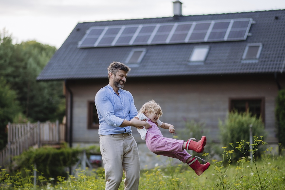 A father and young daughter play in the yard of a home with solar panels on the roof.