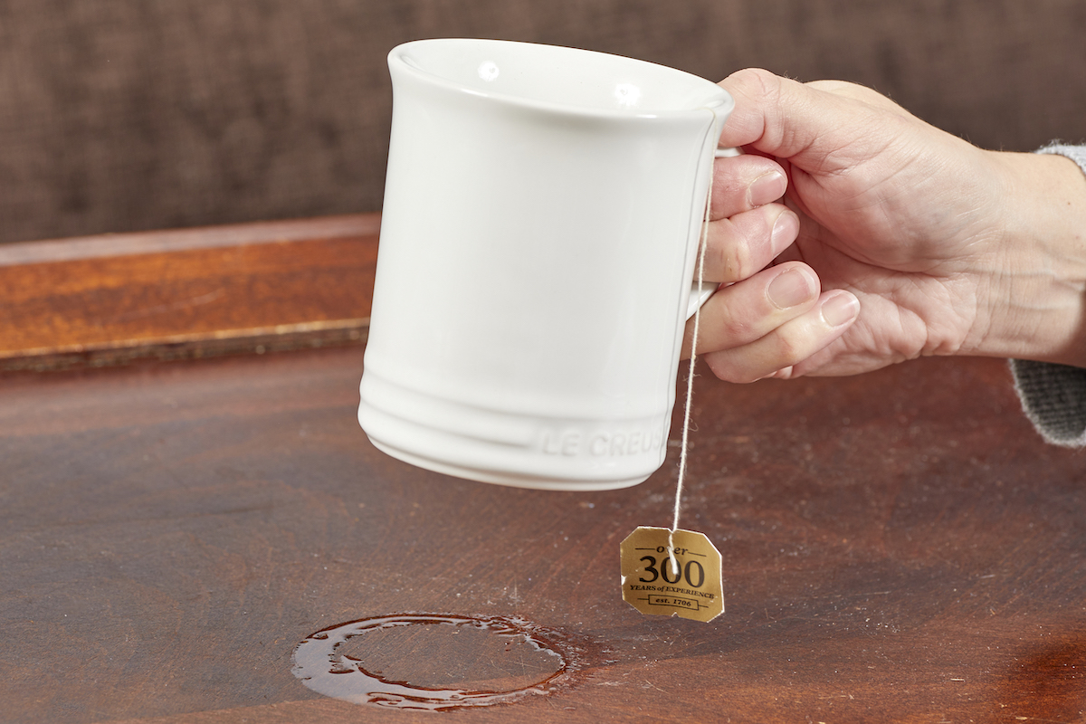 Woman lifts a white teacup off a coffee table to reveal a wet ring on the table.
