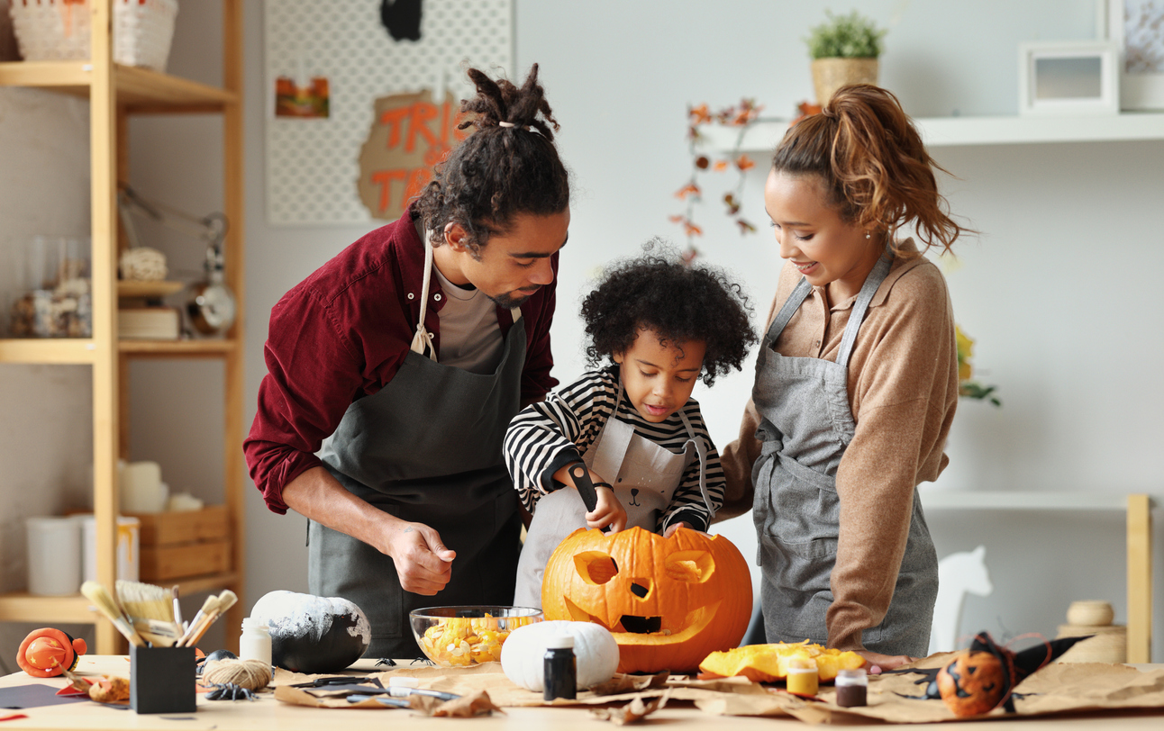 Happy ethnic family mother, father and son carving pumpkin for Halloween holiday together, preparing for holiday party in kitchen, having fun while creating Jack-o-lantern