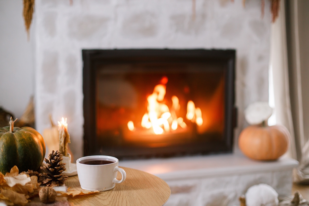 Cozy autumn scene with pumpkins, tea, pinecones in front of a roaring fireplace.