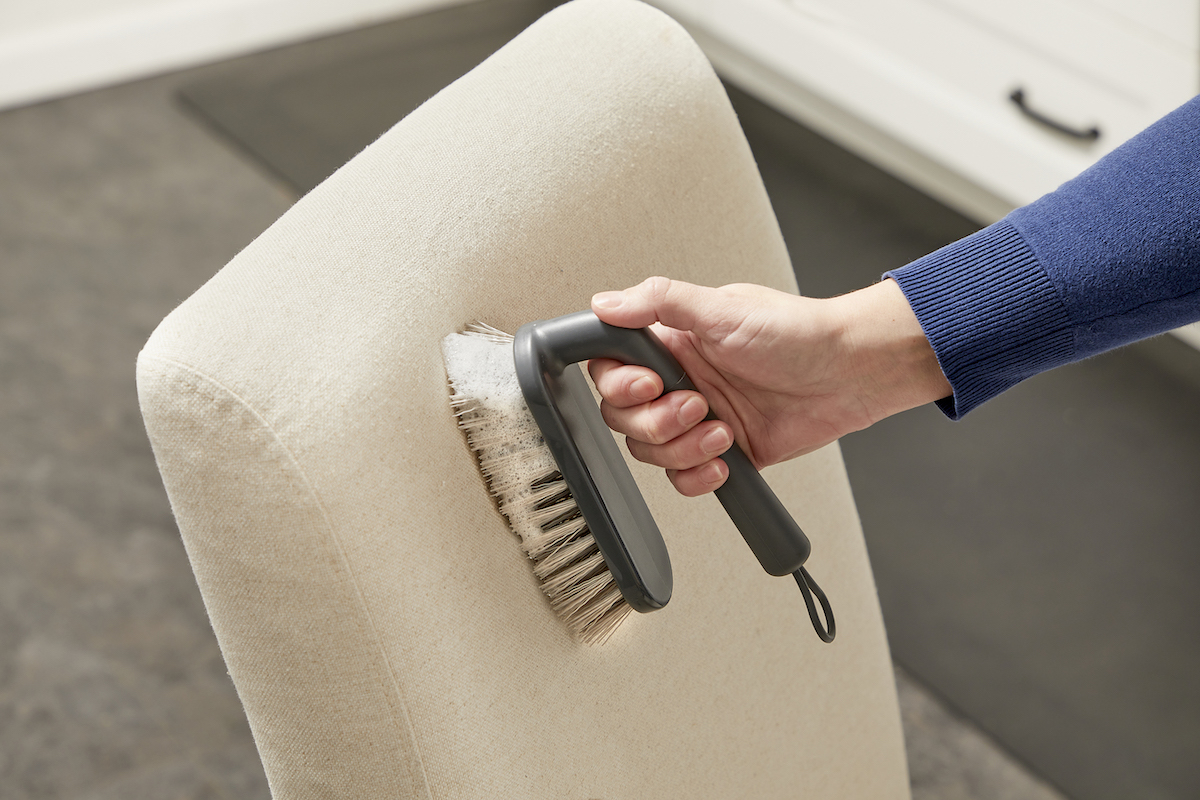 Woman using a sudsy scrub brush to clean a beige upholstered chair.