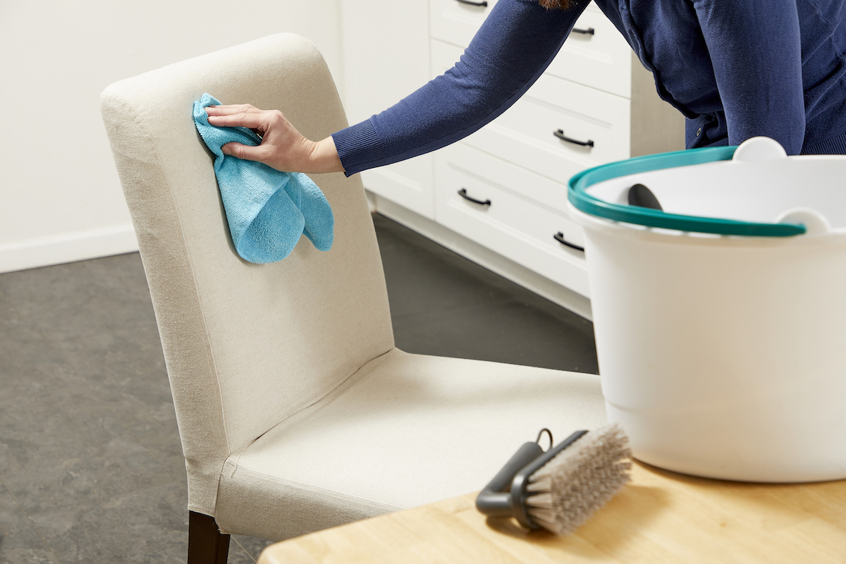 Woman cleaning upholstered chair with a cloth; cleaning supplies in foreground.