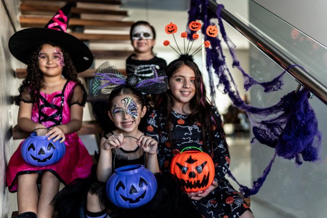 Four young girls wearing Halloween costumes are holding pumpkin-shaped trick-or-treating buckets that are orange or blue.