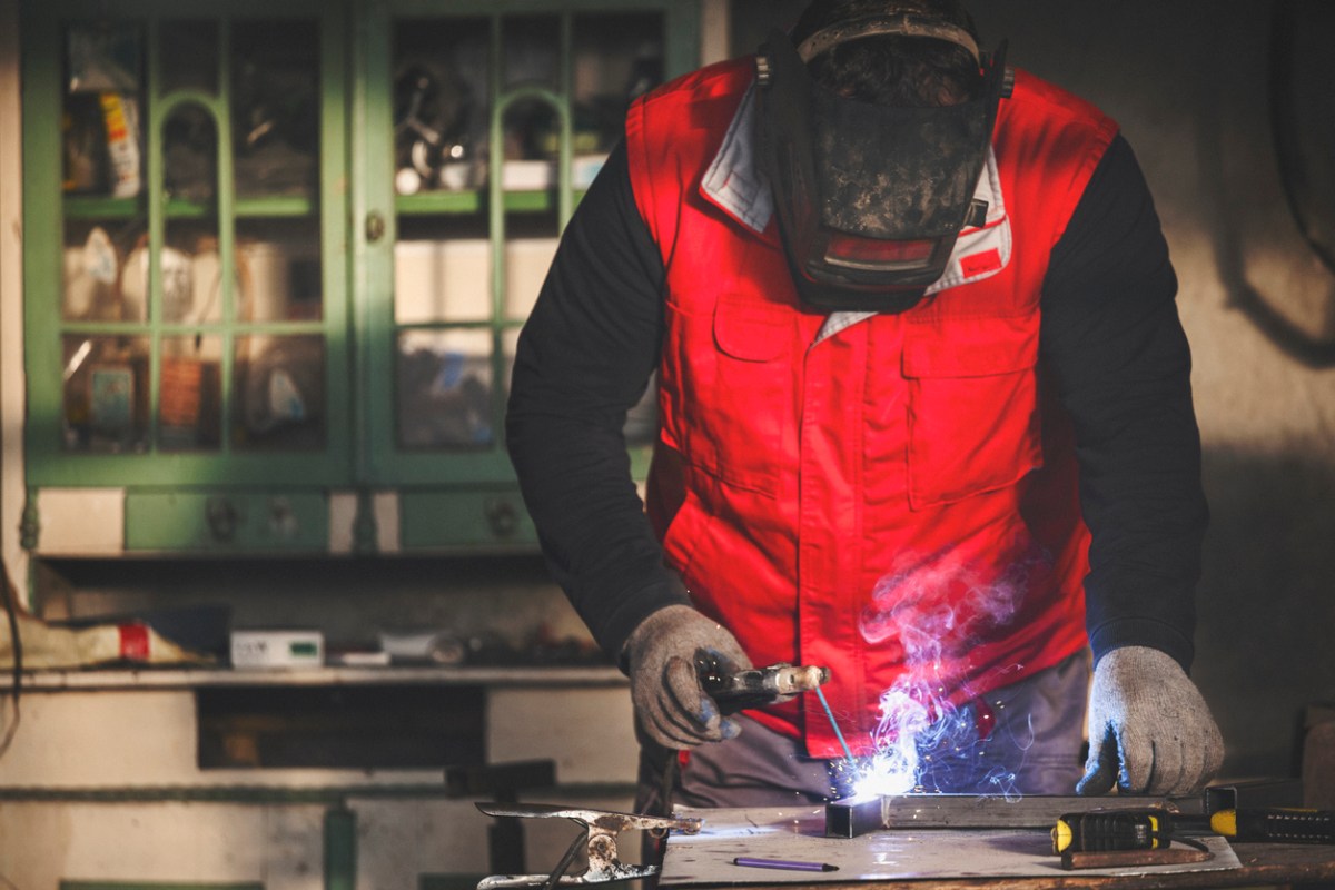 Welder wearing protective equipment and a red vest MIG welding metal in a home workshop