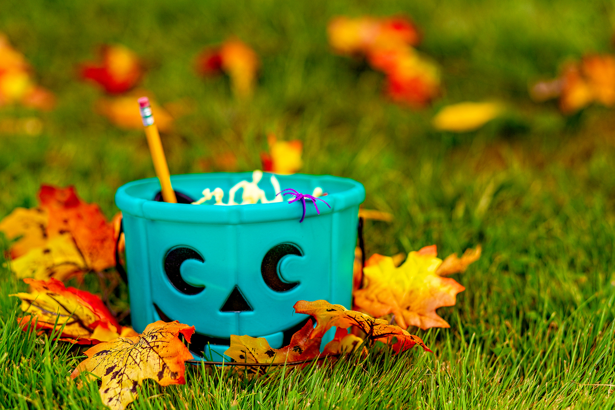 A teal pumpkin candy bucket full of non-food Halloween treats is on grass among orange leaves.