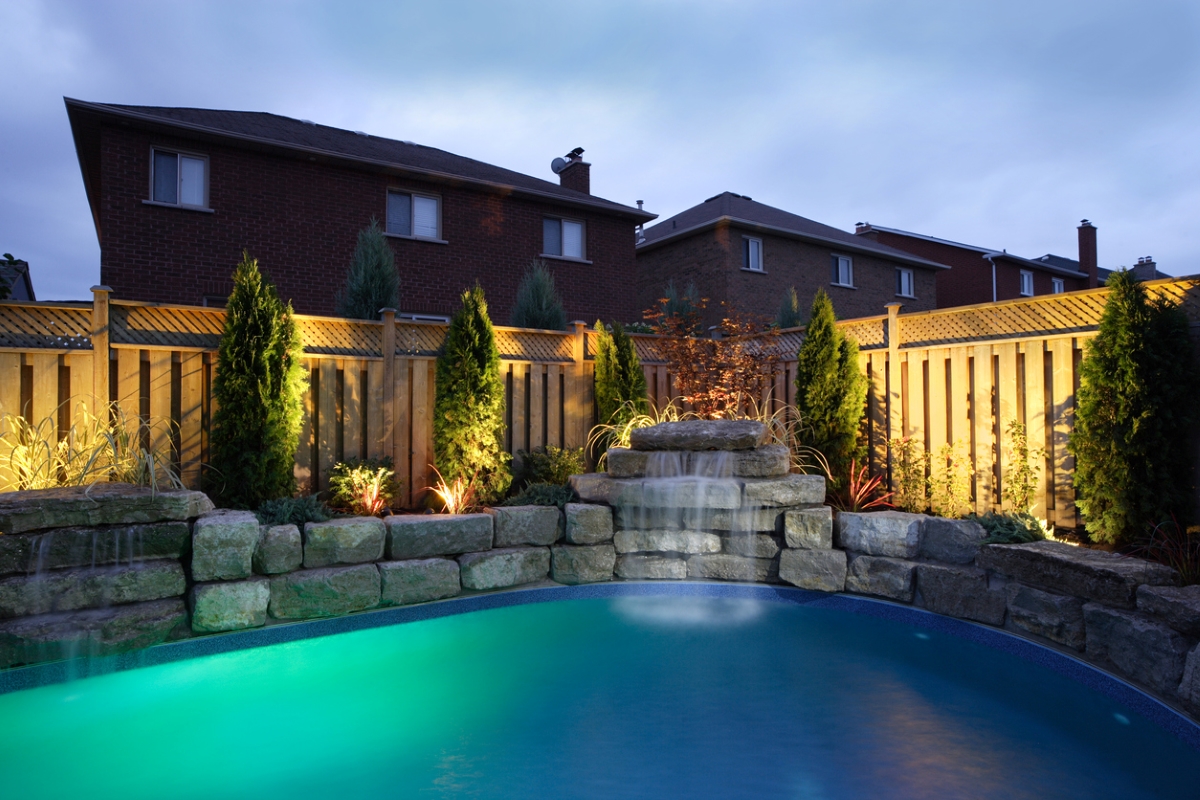 A backyard swimming pool with wooden fence surrounding it and up lighting surrounding its stonework.