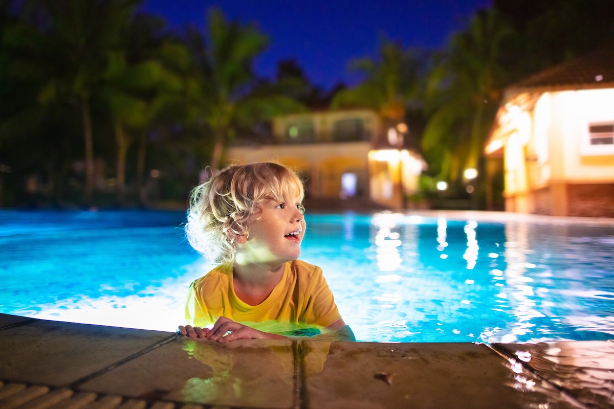 Kid plays in outdoor swimming pool of tropical resort at night.