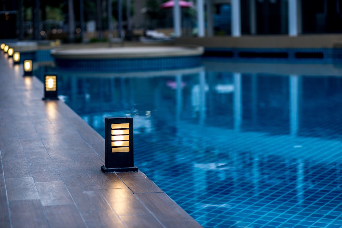 Small black illuminated lanterns line the side of a swimming pool at night.