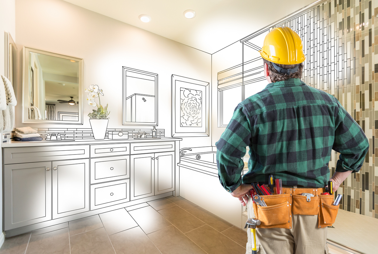 A bathroom remodeling contractor wearing a hard hat and tool belt, looks at a bathroom design drawing overlaid over a plain bathroom.