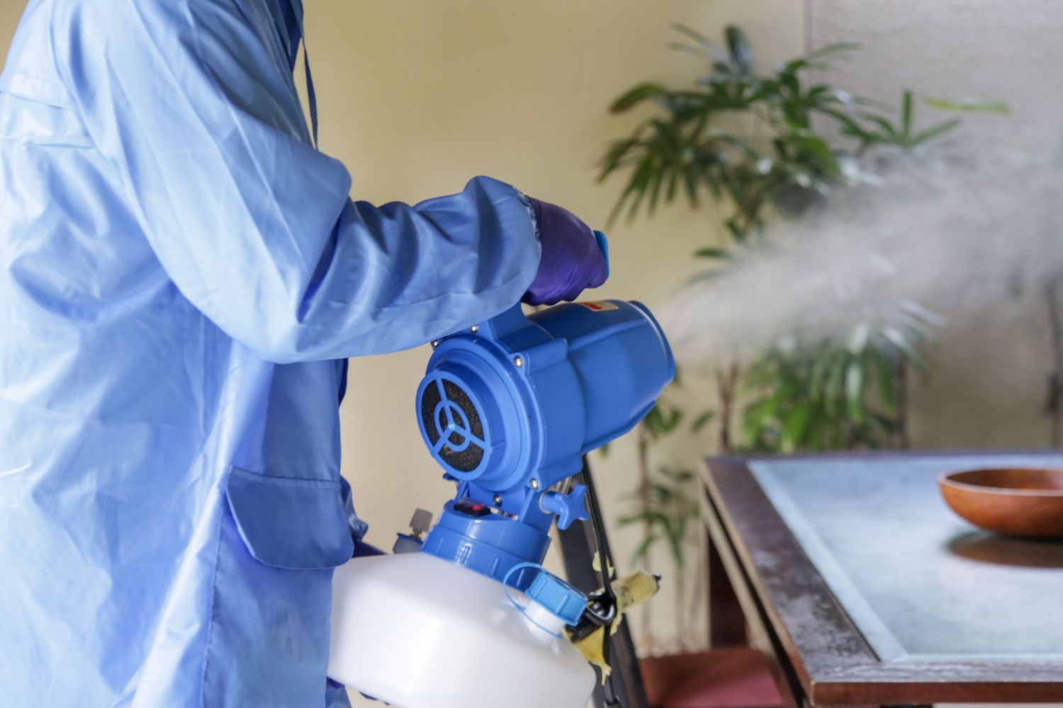 A worker wearing light blue protective clothing is fumigating the inside of a home.