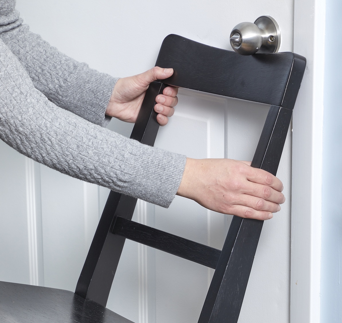 A woman in a gray sweater rests the back of a dark wooden chair under a silver doorknob.