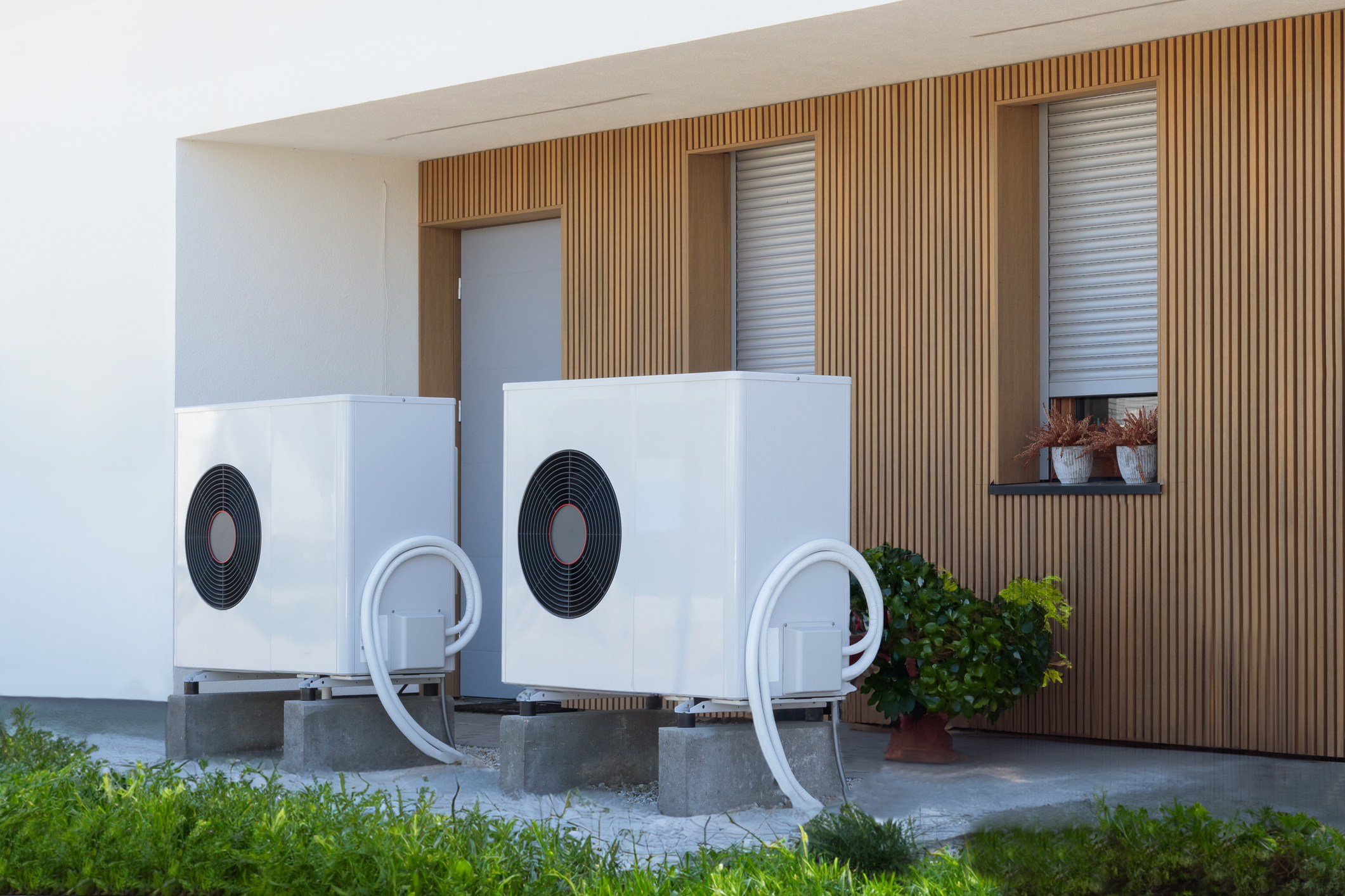 Two white electric heat pumps plugged outdoors and in front of modern home.