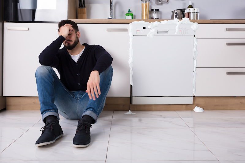 man upset sitting on floor next to foamy dishwasher