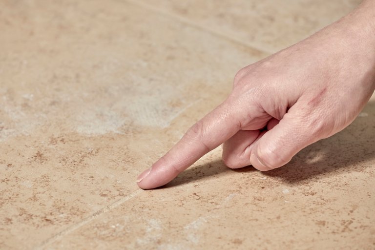 Woman's finger touching grout line between beige tiles; grout haze evident on tiles.