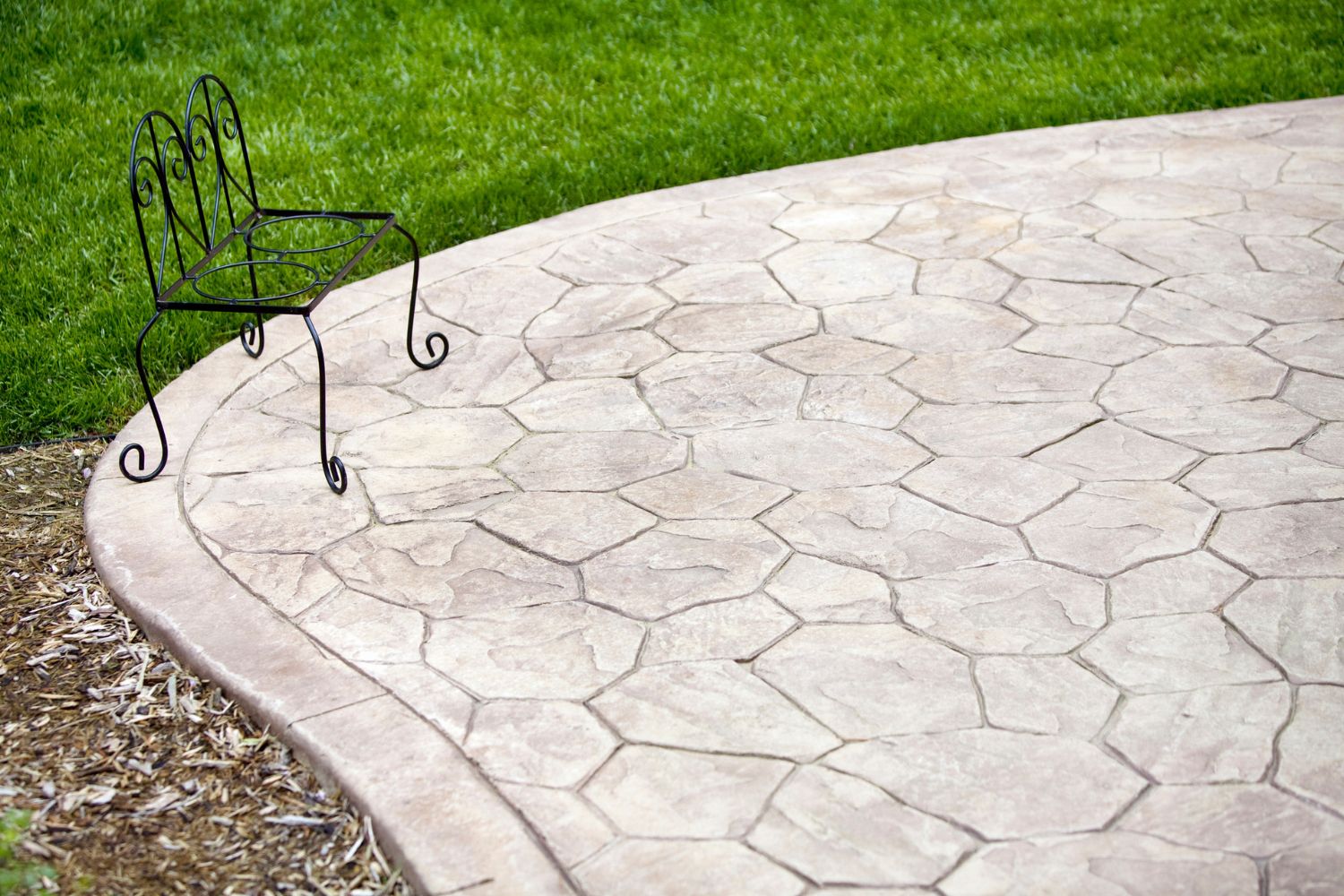 Part of a rounded stamped concrete patio with an iron chair next to a well-manicured lawn.