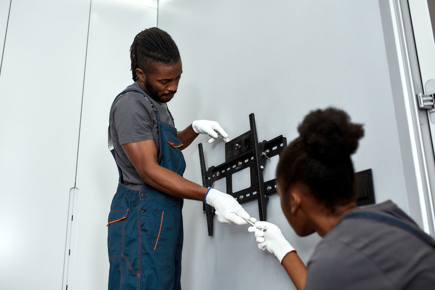 Two workers in overalls install a TV mount on a grey wall. 