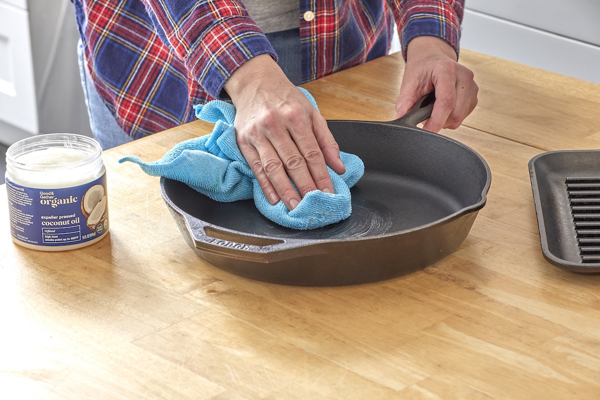 Woman wipes a cast iron skillet with a blue microfiber cloth, a jar of coconut oil nearby.