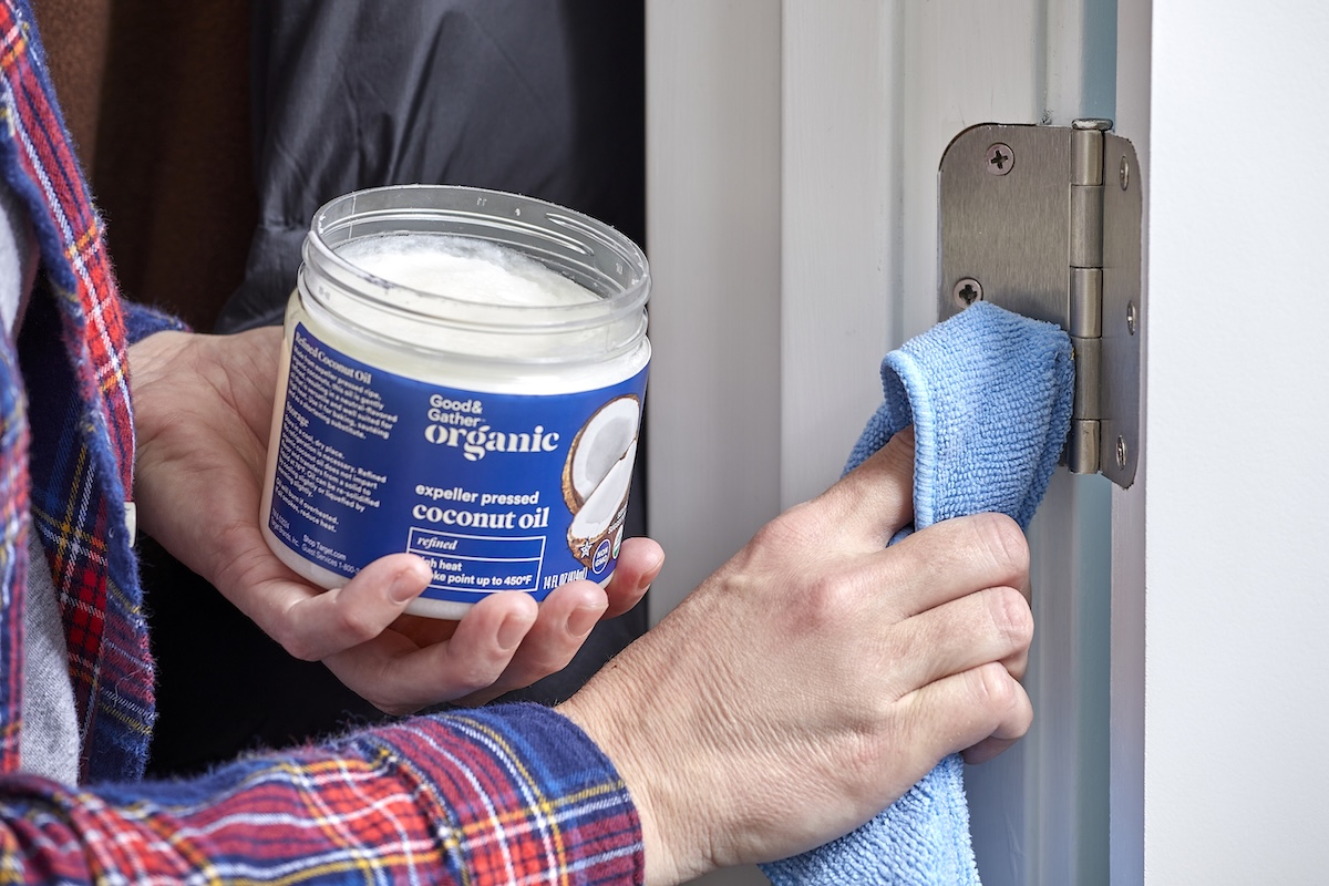 Woman holds jar of coconut oil in one hand and wipes a door hinge with a cloth with the other.