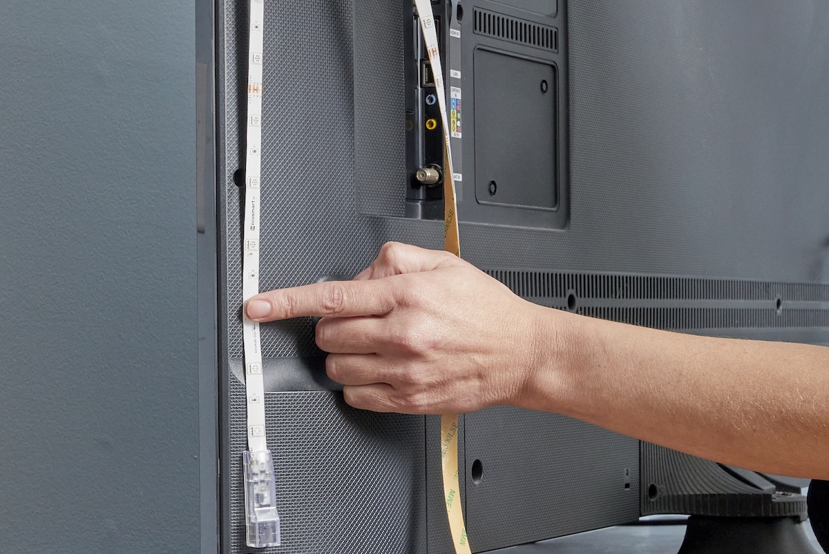 Person affixes beginning of LED light strip to one side of the back of a TV and holds it in place with a finger.