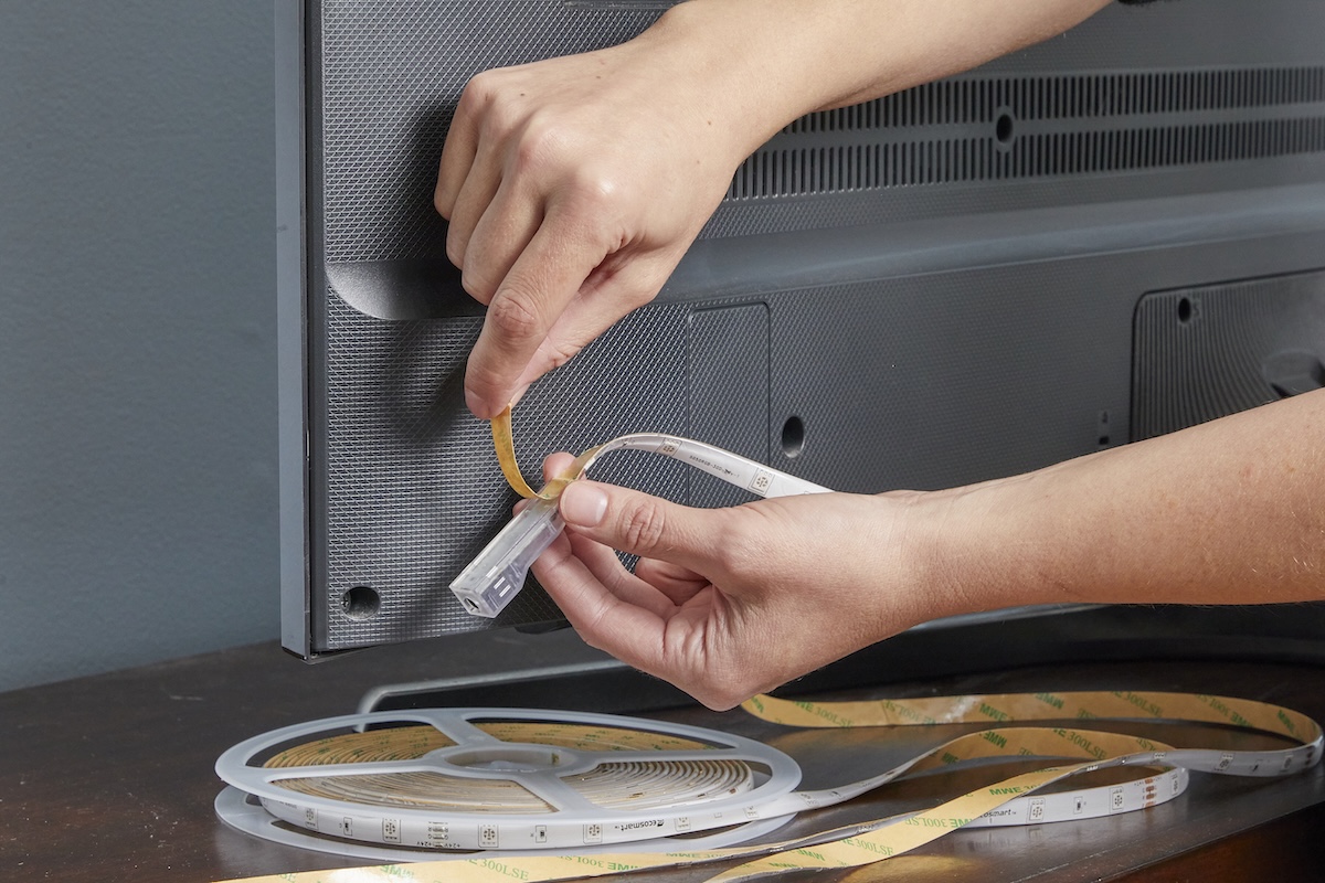Person peels backing off a spool of LED light strips next to the back of a TV.