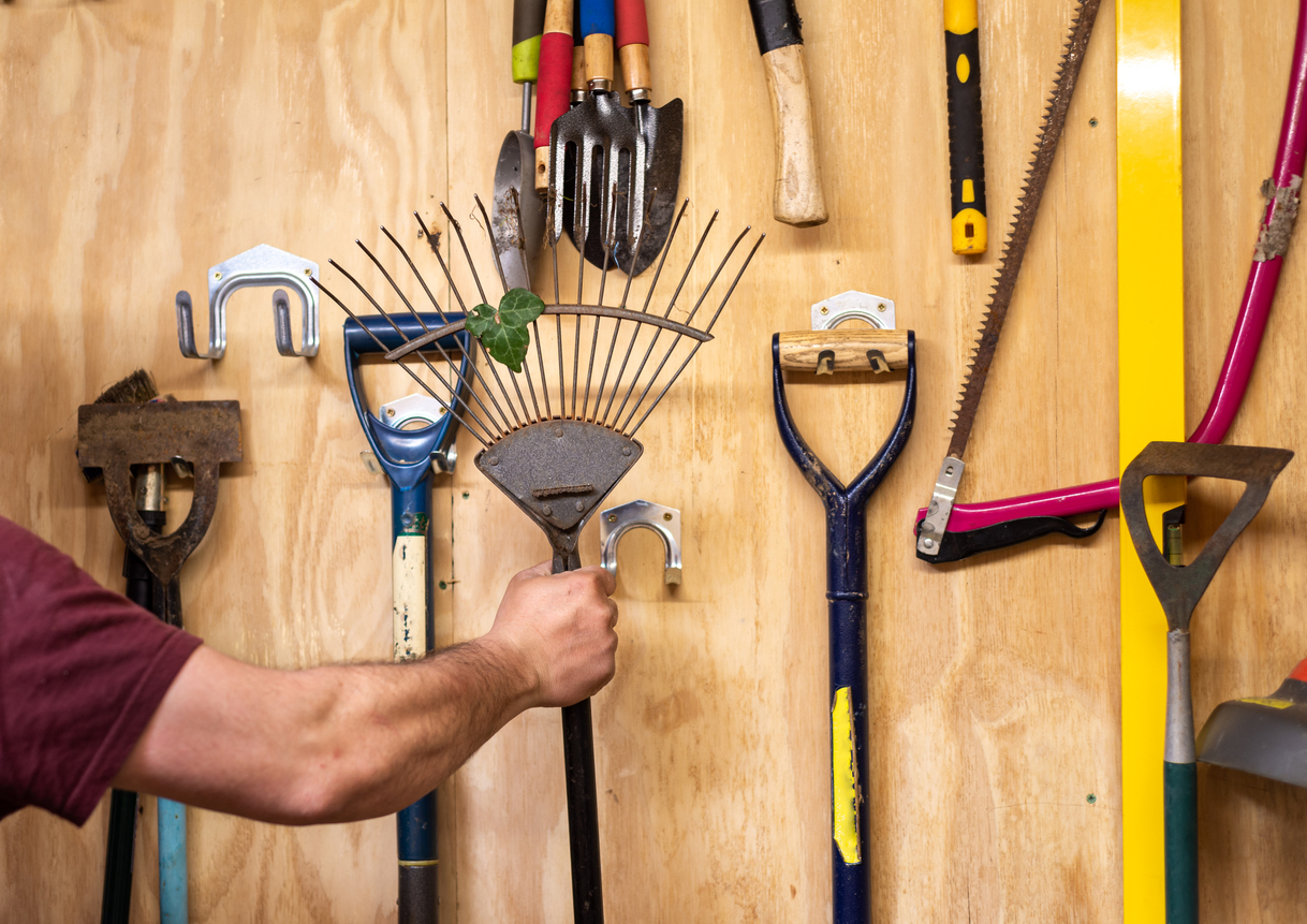 iStock-1434697376 spring cleaning gardening tools Man's arm taking lawn and leaf rake off wooden wall with various hanging DIY garden tools in shed
